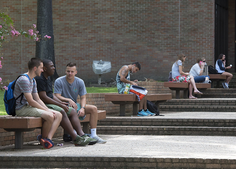 Three students talk on a bench outside an academic building.