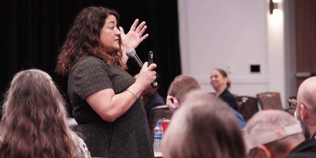 person gesturing while speaking in a mic to a crowd of people