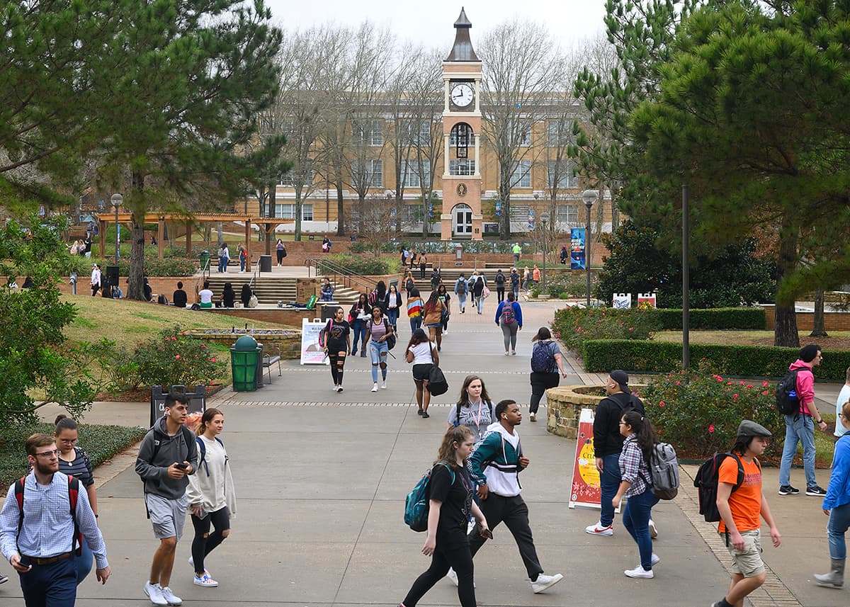 Many SHSU students traversing the middle of campus on a cloudy day. The clocktower stands in the background.