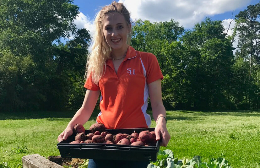 Courtney Rosenbalm kneels in the grass and shows off a tray of root vegetables.