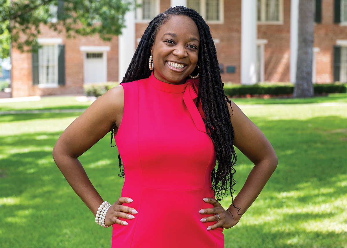 Jeanine Bias poses on the green grass outside of Austin Hall. She is wearing a deep pink dress and has her hands on her hips as she smiles at the camera.